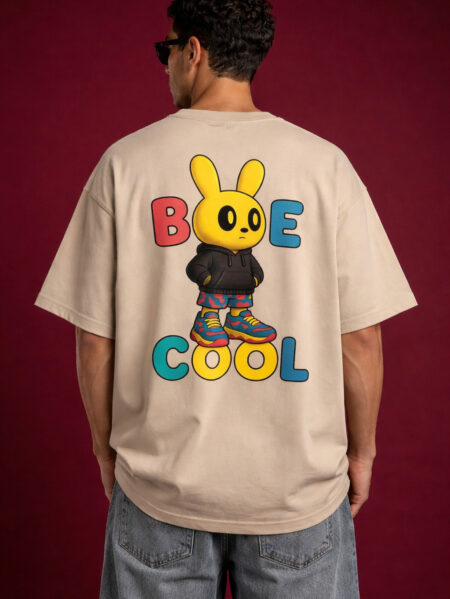 Beige oversized t-shirt worn by male model featuring large colorful “Be Cool” text with cartoon bunny graphic on back, styled with grey jeans on red background