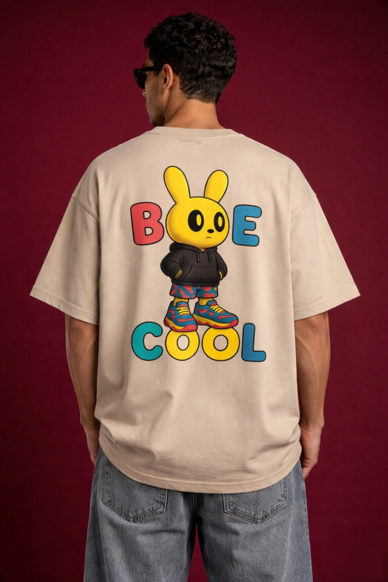 Beige oversized t-shirt worn by male model featuring large colorful “Be Cool” text with cartoon bunny graphic on back, styled with grey jeans on red background