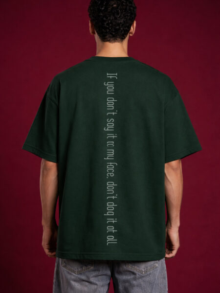Back view of a boxy fit dark green streetwear t-shirt showing a vertical spine-aligned text graphic that reads 'If you don't say it to my face, don't dog it at all.' Minimalist streetwear design with drop shoulder construction on a male model.