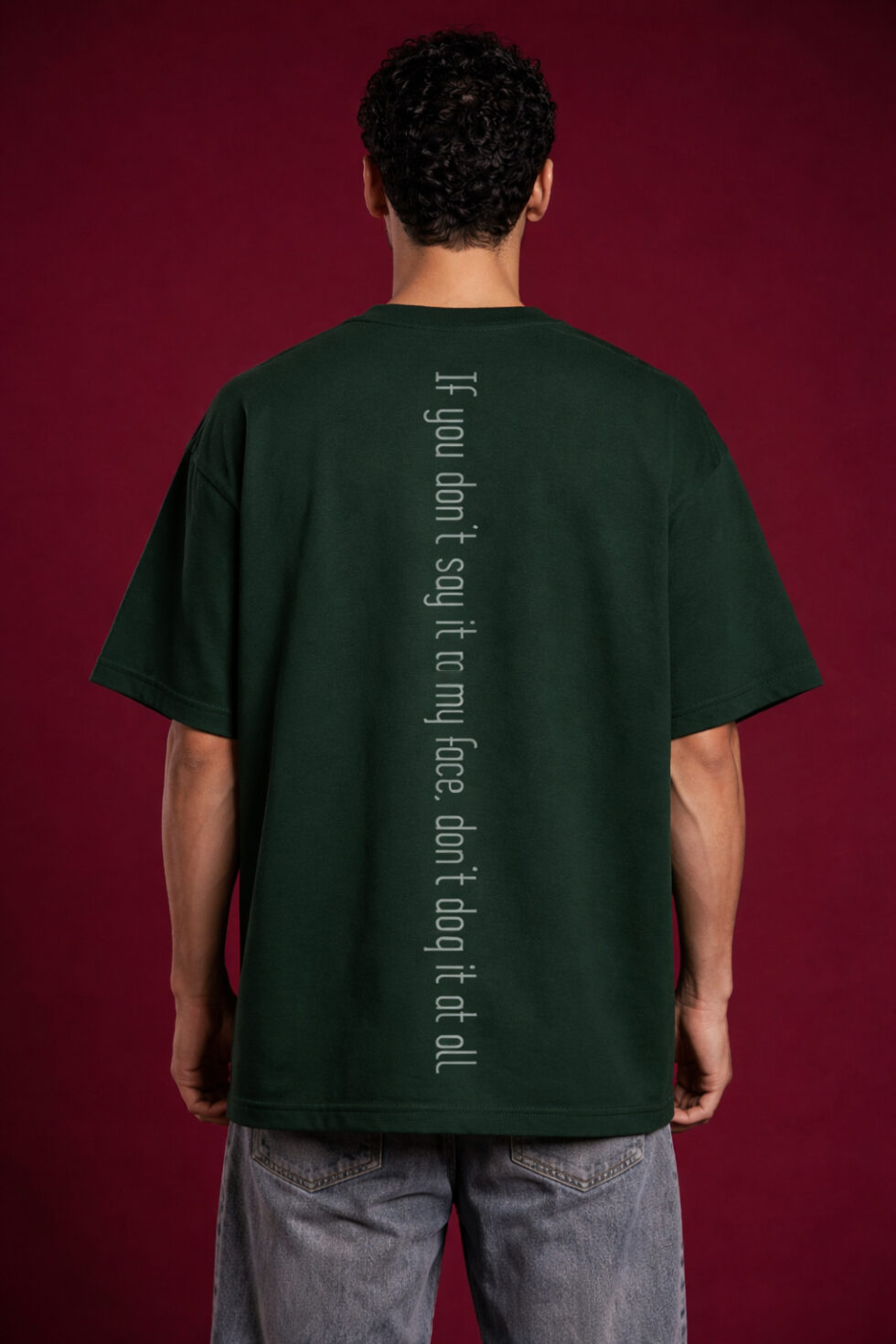 Back view of a boxy fit dark green streetwear t-shirt showing a vertical spine-aligned text graphic that reads 'If you don't say it to my face, don't dog it at all.' Minimalist streetwear design with drop shoulder construction on a male model.