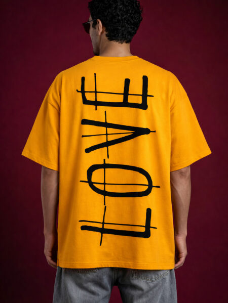 Back view of a mustard yellow drop-shoulder oversized t-shirt featuring a vertical, minimalist "LOVE" typography design in a bold, distressed black ink-wash style. The streetwear fit is paired with grey denim for a modern grunge aesthetic.