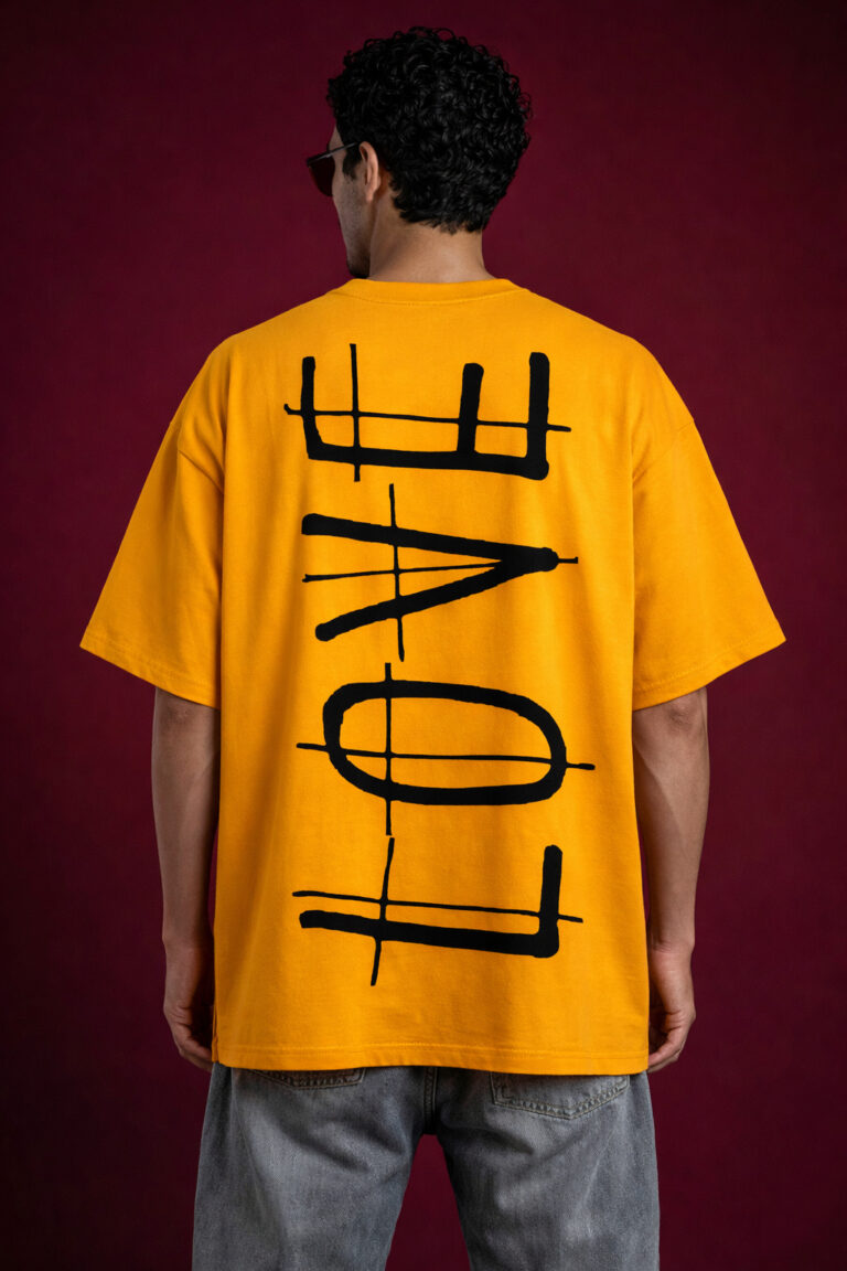 Back view of a mustard yellow drop-shoulder oversized t-shirt featuring a vertical, minimalist "LOVE" typography design in a bold, distressed black ink-wash style. The streetwear fit is paired with grey denim for a modern grunge aesthetic.