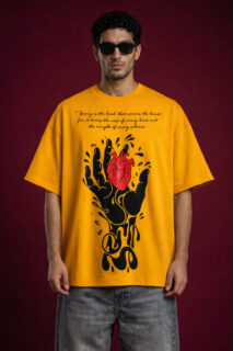 Male model wearing a mustard yellow oversized streetwear t-shirt featuring a large graphic of a black melting hand holding a realistic red heart. The chest includes elegant cursive typography and the garment is made from heavyweight cotton fleece with a boxy fit.