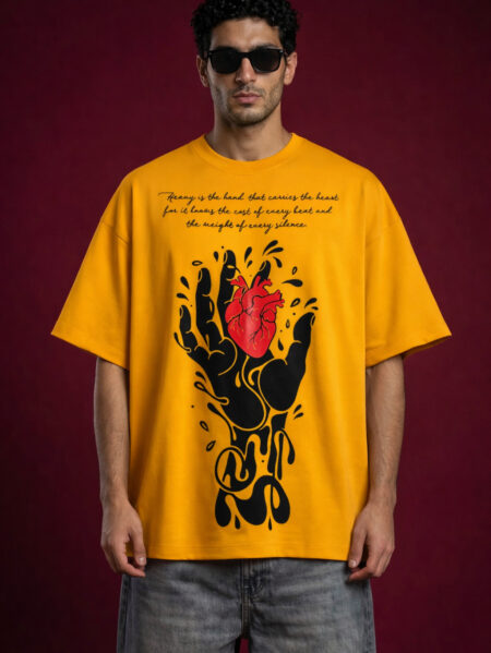 Male model wearing a mustard yellow oversized streetwear t-shirt featuring a large graphic of a black melting hand holding a realistic red heart. The chest includes elegant cursive typography and the garment is made from heavyweight cotton fleece with a boxy fit.