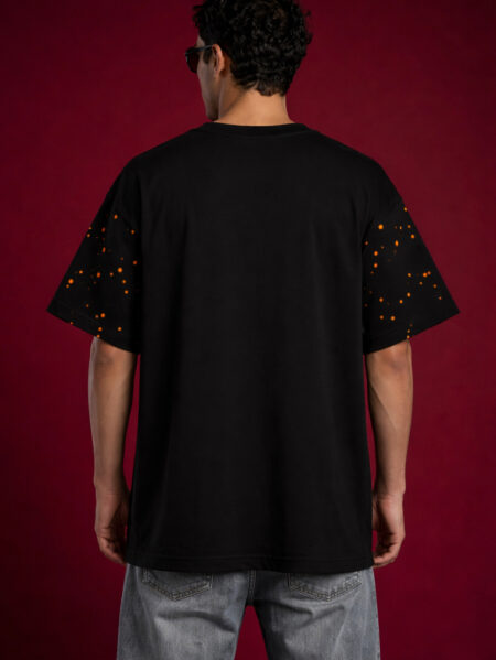 Back view of a black heavyweight oversized t-shirt on a male model. The design highlights an orange speckled paint splatter pattern on the short sleeves and a relaxed, boxy fit against a dark red background.