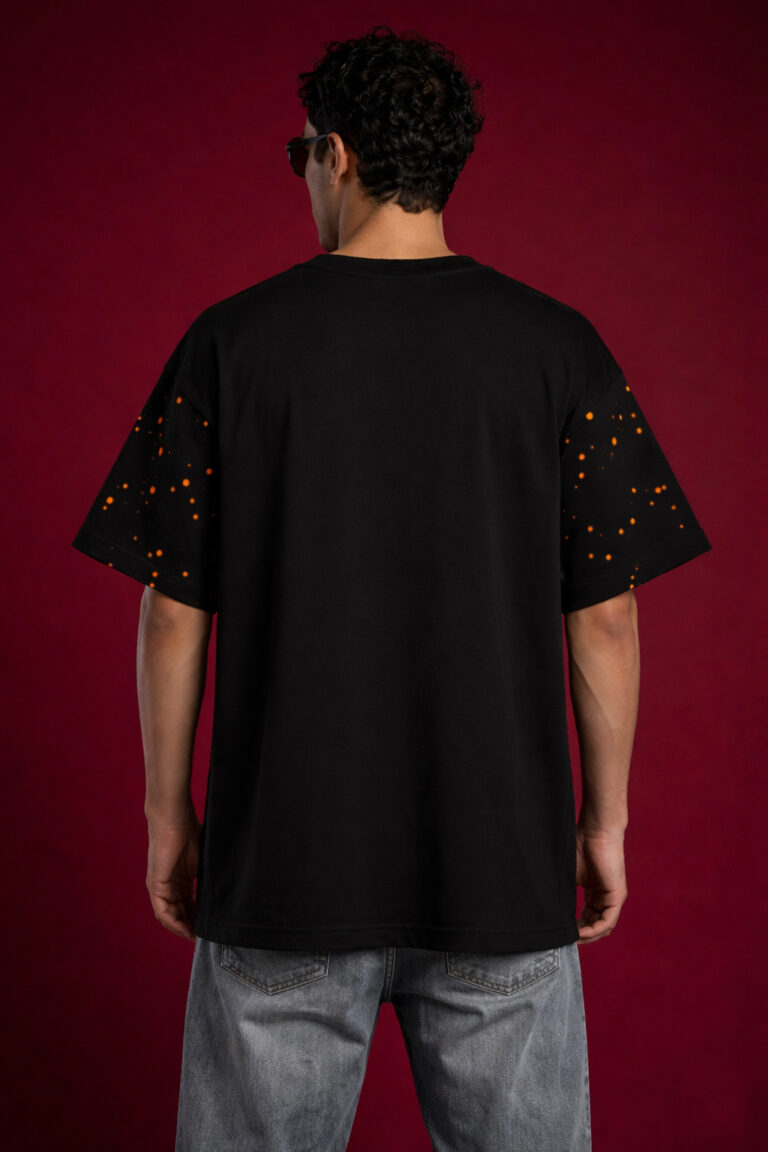 Back view of a black heavyweight oversized t-shirt on a male model. The design highlights an orange speckled paint splatter pattern on the short sleeves and a relaxed, boxy fit against a dark red background.