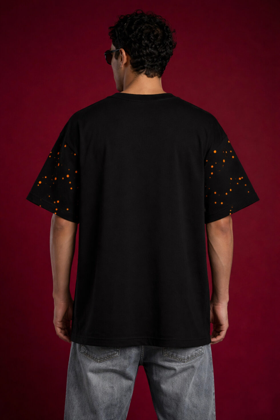 Back view of a black heavyweight oversized t-shirt on a male model. The design highlights an orange speckled paint splatter pattern on the short sleeves and a relaxed, boxy fit against a dark red background.