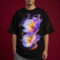Male model wearing a black oversized streetwear t-shirt featuring a vibrant purple and blue floral graphic with orange swirl accents. The t-shirt has dropped shoulders and orange paint splatter details on the sleeves