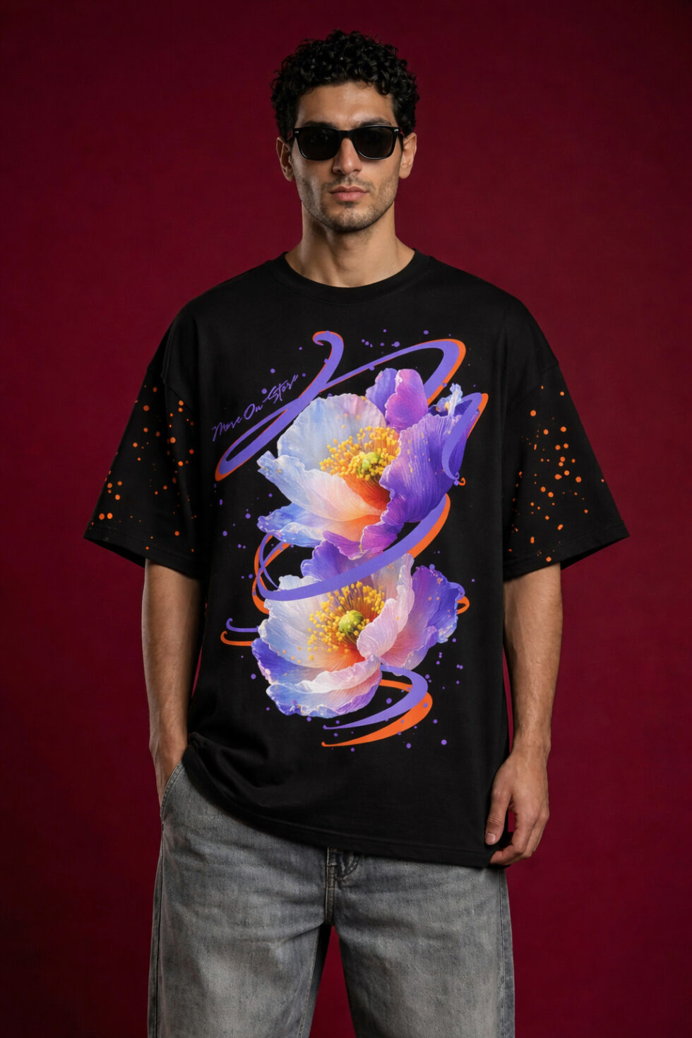 Male model wearing a black oversized streetwear t-shirt featuring a vibrant purple and blue floral graphic with orange swirl accents. The t-shirt has dropped shoulders and orange paint splatter details on the sleeves