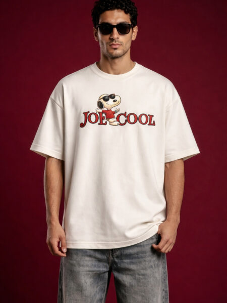 Front view of a man wearing a cream-colored oversized streetwear t-shirt featuring a 'Joe Cool' Snoopy graphic and red typography on the chest. The model is wearing black sunglasses and standing against a dark maroon background.