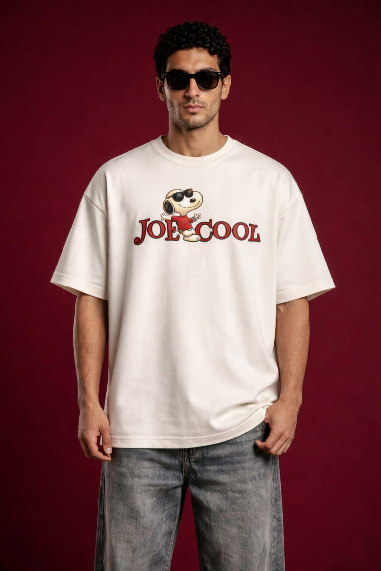 Front view of a man wearing a cream-colored oversized streetwear t-shirt featuring a 'Joe Cool' Snoopy graphic and red typography on the chest. The model is wearing black sunglasses and standing against a dark maroon background.