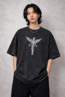 A male model wearing a premium oversized acid-wash charcoal t-shirt. The shirt features a centered white graphic of a skeletal winged angel in a 90s grunge style. The fit is boxy with drop-shoulder sleeves, styled with black trousers against a neutral industrial concrete background.