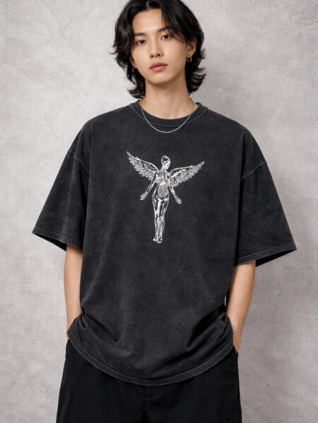 A male model wearing a premium oversized acid-wash charcoal t-shirt. The shirt features a centered white graphic of a skeletal winged angel in a 90s grunge style. The fit is boxy with drop-shoulder sleeves, styled with black trousers against a neutral industrial concrete background.