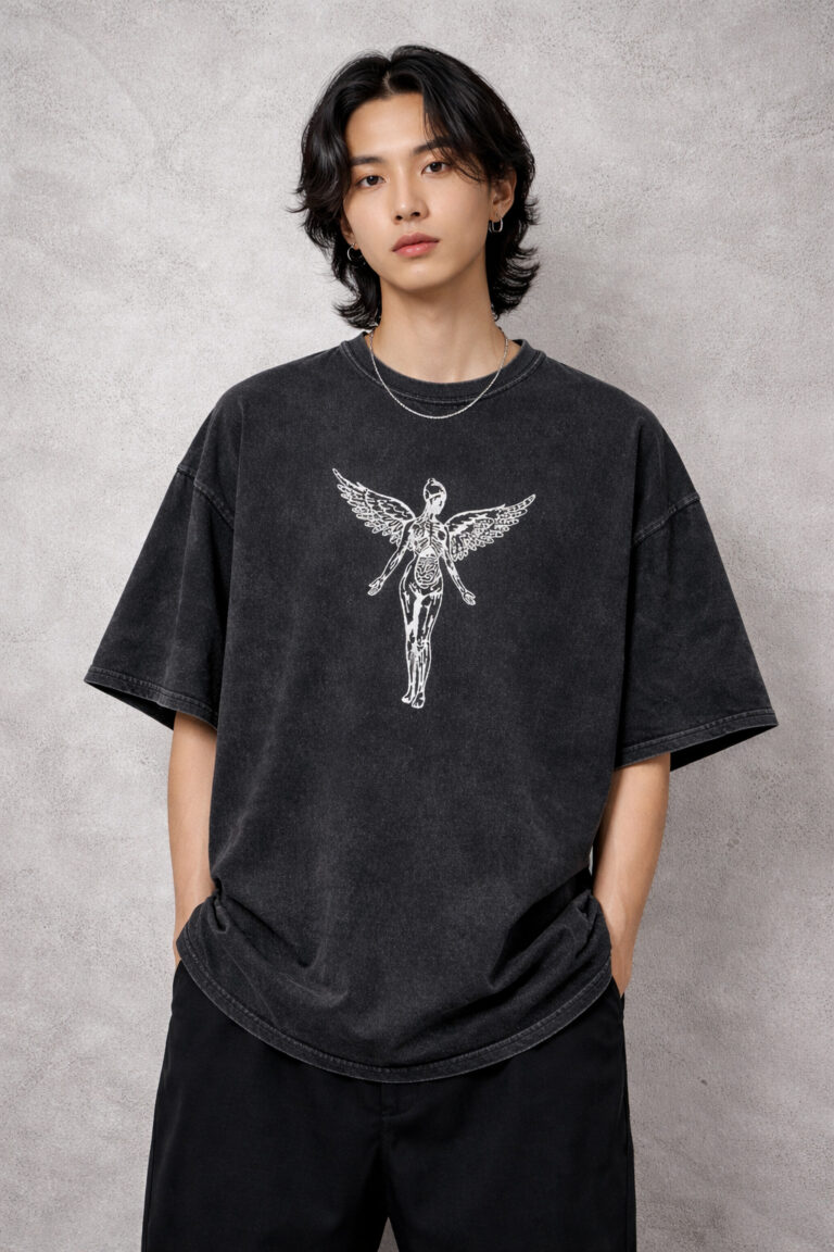 A male model wearing a premium oversized acid-wash charcoal t-shirt. The shirt features a centered white graphic of a skeletal winged angel in a 90s grunge style. The fit is boxy with drop-shoulder sleeves, styled with black trousers against a neutral industrial concrete background.