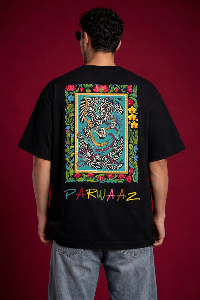 Back view of a black oversized graphic t-shirt featuring a large, vibrant South Asian truck art-inspired phoenix illustration inside a floral border, with "PARWAAZ" text printed in colorful hand-drawn font at the bottom.