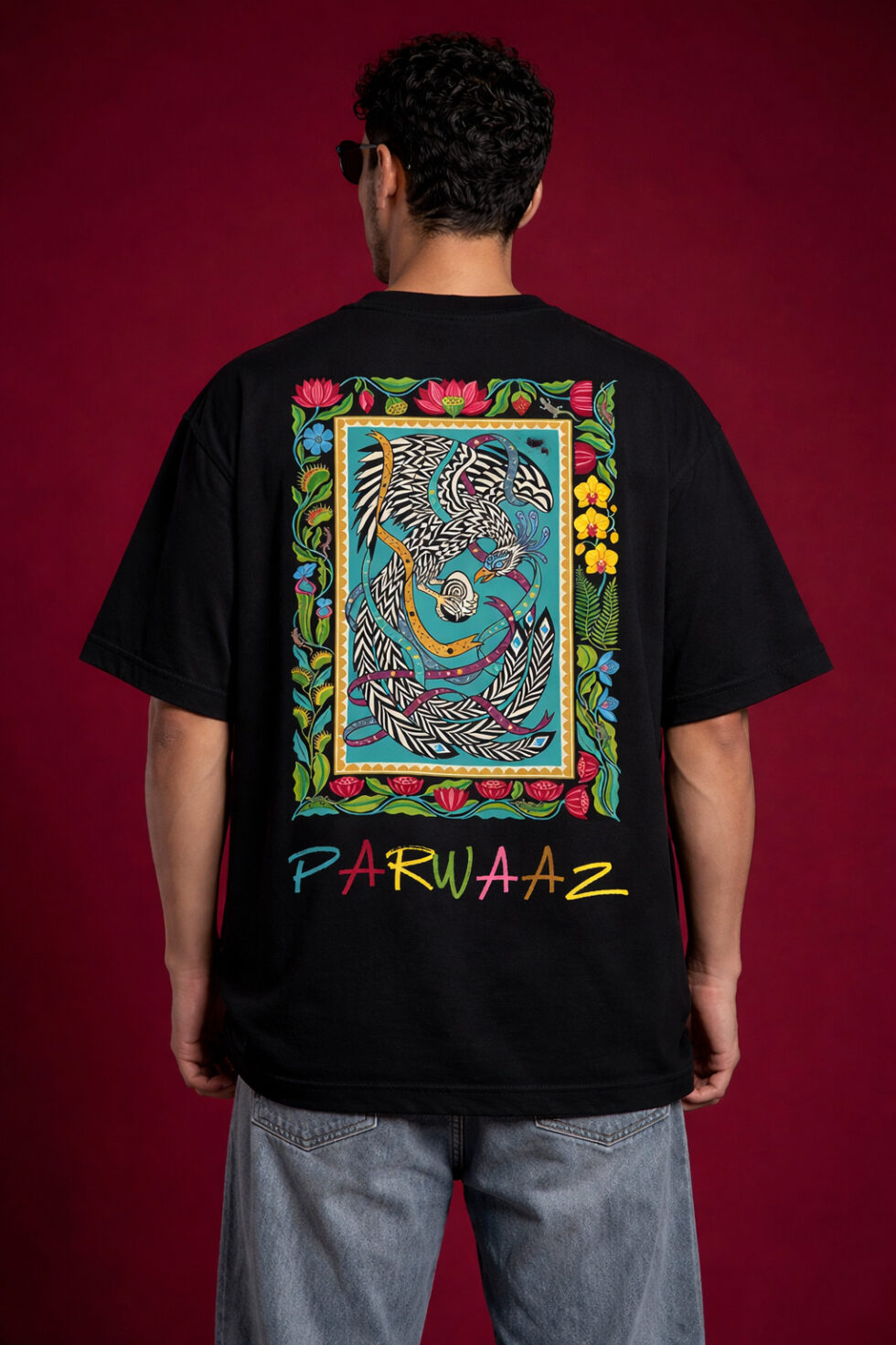 Back view of a black oversized graphic t-shirt featuring a large, vibrant South Asian truck art-inspired phoenix illustration inside a floral border, with "PARWAAZ" text printed in colorful hand-drawn font at the bottom.