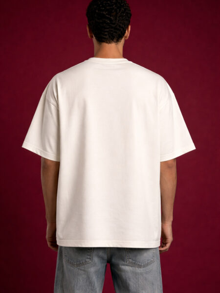 Back view of a man wearing a premium off-white heavyweight oversized t-shirt. The image highlights the clean, minimalist back, dropped shoulder seams, and relaxed boxy silhouette. The model is set against a textured dark red backdrop.