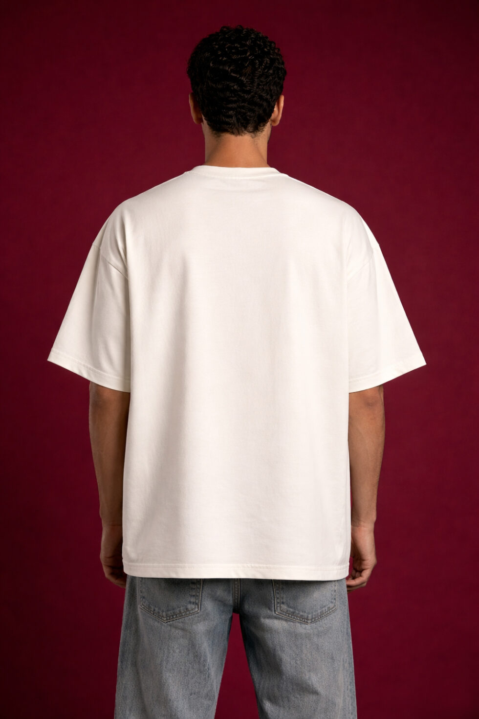 Back view of a man wearing a premium off-white heavyweight oversized t-shirt. The image highlights the clean, minimalist back, dropped shoulder seams, and relaxed boxy silhouette. The model is set against a textured dark red backdrop.