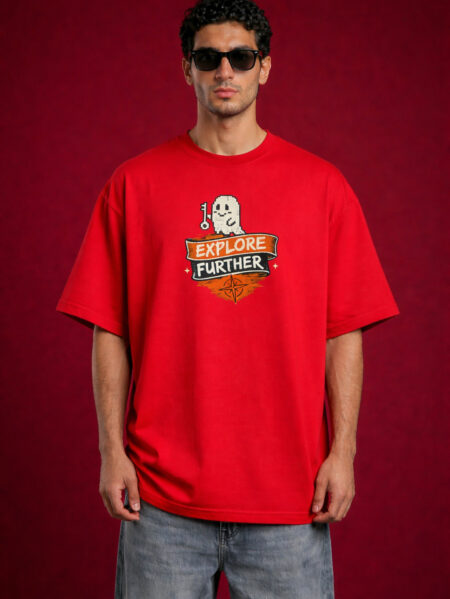 A male model wearing a bright red, oversized streetwear t-shirt. The chest features a graphic of a white pixel-art ghost holding a key above a banner that says "EXPLORE FURTHER" with a compass icon. The model is also wearing dark sunglasses and light-wash denim jeans against a deep red textured background.