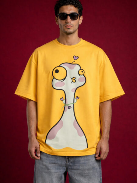 Front view of a male model wearing an oversized mustard yellow graphic t-shirt featuring a large whimsical cartoon character with pink spots and purple hearts.