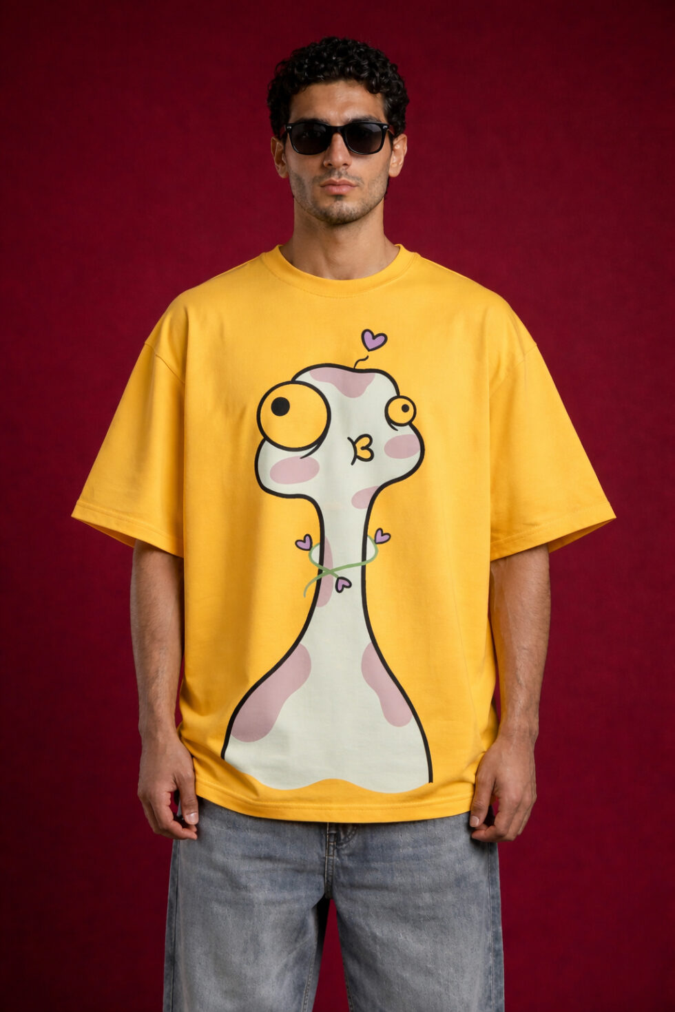 Front view of a male model wearing an oversized mustard yellow graphic t-shirt featuring a large whimsical cartoon character with pink spots and purple hearts.