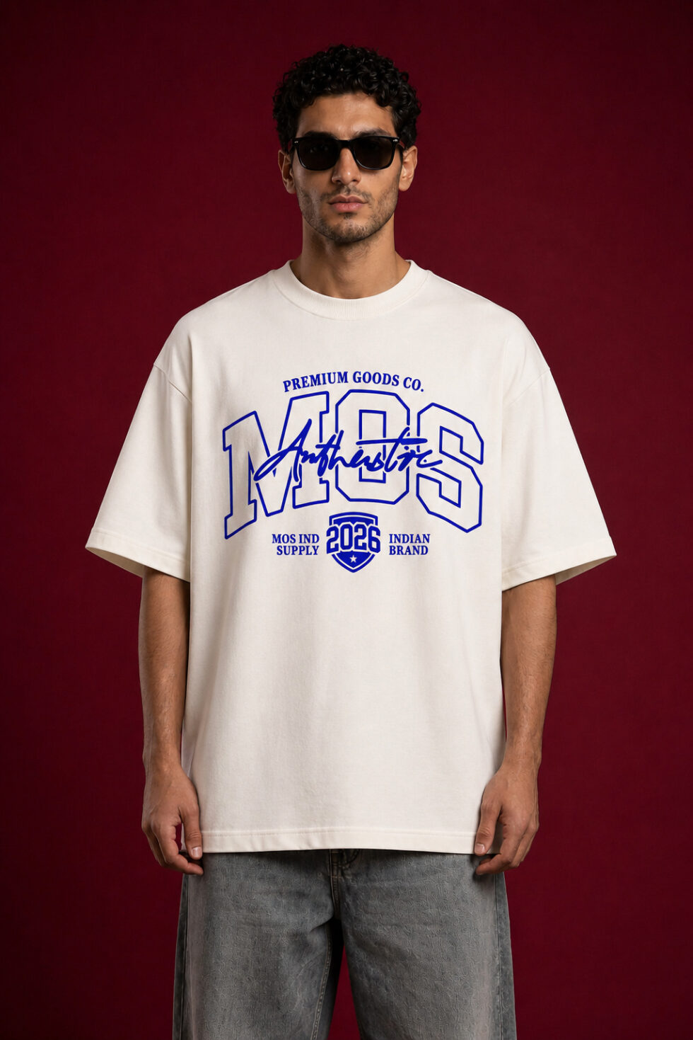 A man wearing an oversized off-white streetwear t-shirt with a bold blue "MOS" graphic on the chest. The design includes "Authentic" in script and a "2026" shield emblem. He is wearing black sunglasses and grey washed denim, standing against a dark maroon textured background