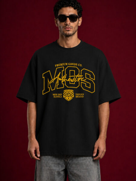 A man with dark curly hair and sunglasses wearing a black oversized t-shirt with a large yellow graphic design. The graphic features the letters "MOS" in a collegiate font with "Authentic" scripted over it and "2026" inside a shield emblem at the bottom.