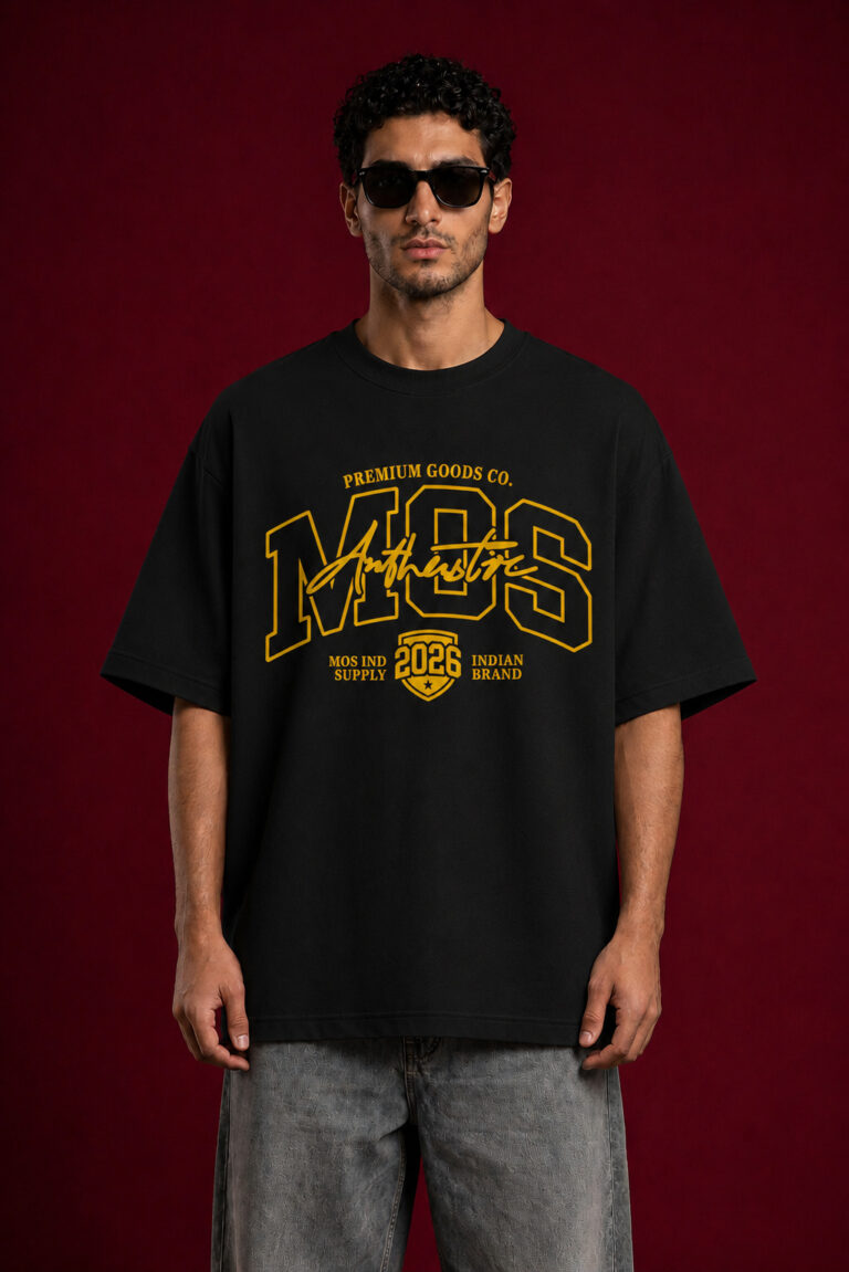 A man with dark curly hair and sunglasses wearing a black oversized t-shirt with a large yellow graphic design. The graphic features the letters "MOS" in a collegiate font with "Authentic" scripted over it and "2026" inside a shield emblem at the bottom.