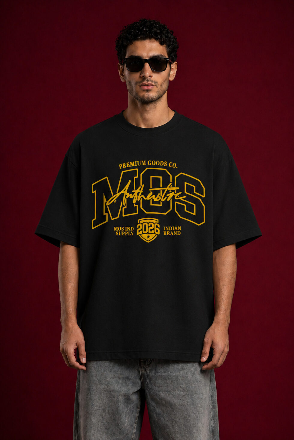 A man with dark curly hair and sunglasses wearing a black oversized t-shirt with a large yellow graphic design. The graphic features the letters "MOS" in a collegiate font with "Authentic" scripted over it and "2026" inside a shield emblem at the bottom.
