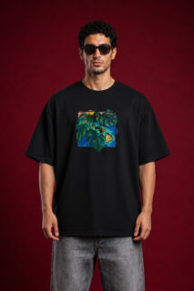 Full-length front view of a man with curly hair wearing sunglasses and a black oversized graphic t-shirt. The shirt features a centered, square-framed colorful illustration of large green monstera leaves layered over a blue and yellow tropical island map design. He is wearing faded gray jeans against a solid dark red background