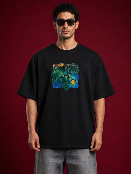 Full-length front view of a man with curly hair wearing sunglasses and a black oversized graphic t-shirt. The shirt features a centered, square-framed colorful illustration of large green monstera leaves layered over a blue and yellow tropical island map design. He is wearing faded gray jeans against a solid dark red background