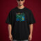 Full-length front view of a man with curly hair wearing sunglasses and a black oversized graphic t-shirt. The shirt features a centered, square-framed colorful illustration of large green monstera leaves layered over a blue and yellow tropical island map design. He is wearing faded gray jeans against a solid dark red background
