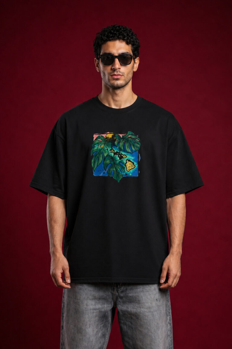 Full-length front view of a man with curly hair wearing sunglasses and a black oversized graphic t-shirt. The shirt features a centered, square-framed colorful illustration of large green monstera leaves layered over a blue and yellow tropical island map design. He is wearing faded gray jeans against a solid dark red background