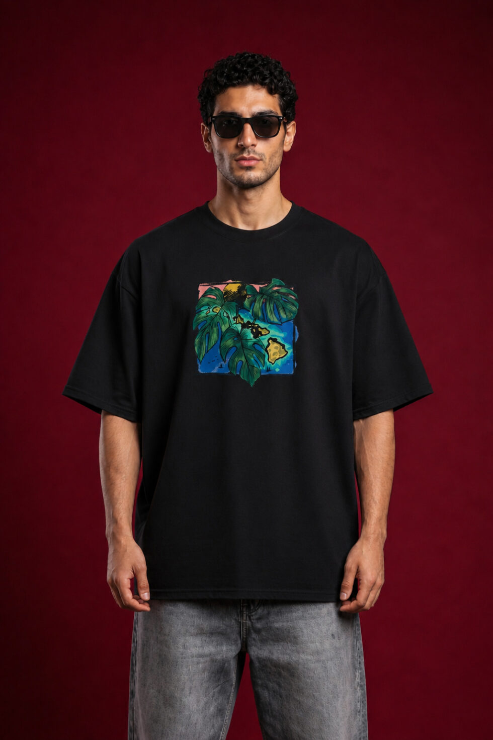 Full-length front view of a man with curly hair wearing sunglasses and a black oversized graphic t-shirt. The shirt features a centered, square-framed colorful illustration of large green monstera leaves layered over a blue and yellow tropical island map design. He is wearing faded gray jeans against a solid dark red background