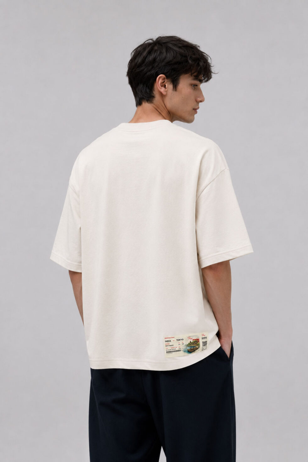 Back view of a man in a loose-fitting cream streetwear t-shirt with a minimalist design, highlighting a detailed rectangular travel ticket patch stitched near the bottom corner hem.