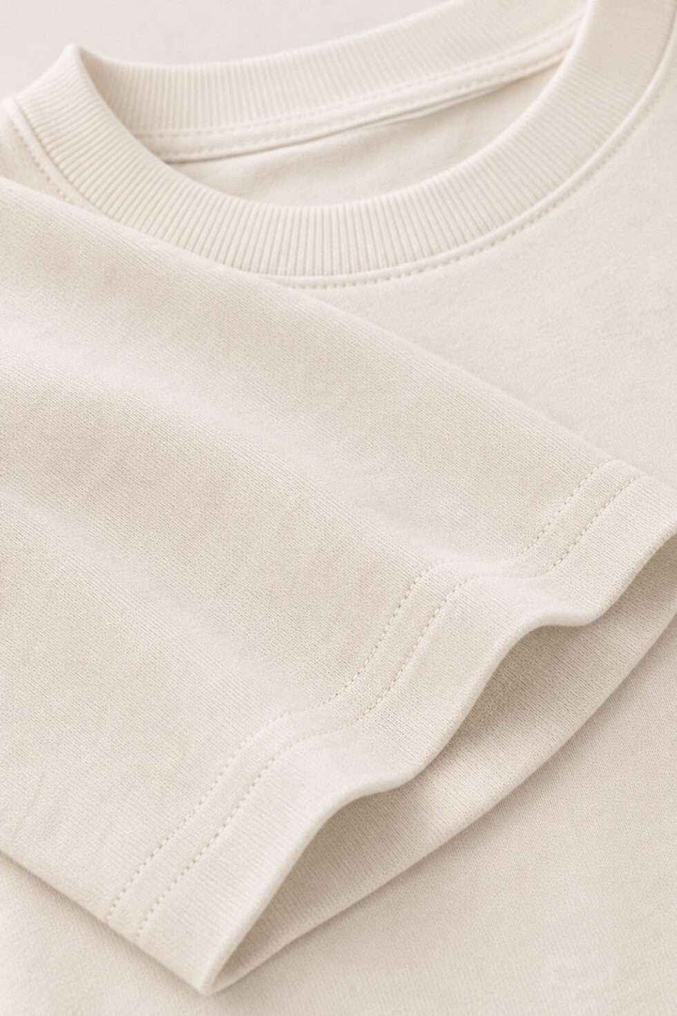 Close-up texture shot of a high-quality off-white heavyweight cotton t-shirt, showing the durable ribbed crewneck neckline and neat double-stitching details on the folded sleeve hem.