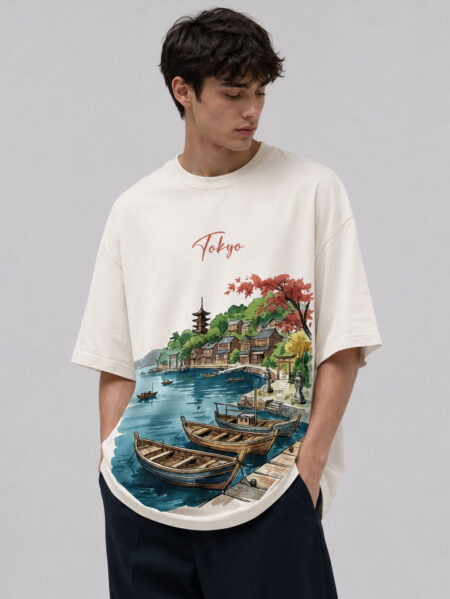 Front view of a man wearing a casual off-white oversized graphic tee featuring a colorful watercolor illustration of a traditional Japanese coastal village with wooden boats, a pagoda, and red autumn trees under a cursive "Tokyo" text.