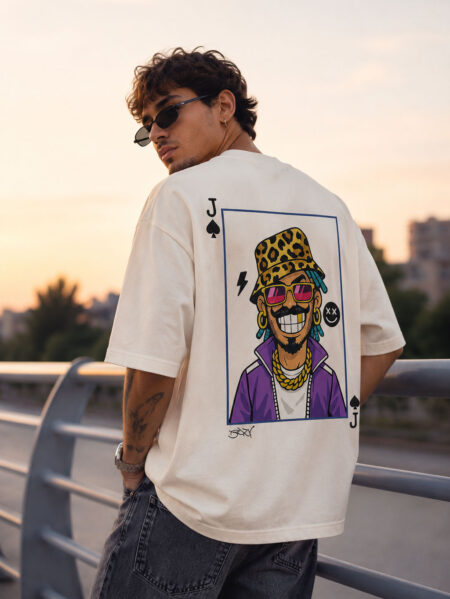 Back view of a man wearing a cream oversized streetwear t-shirt featuring a large cartoon graphic of a character with a gold tooth, leopard bucket hat, and purple jacket inside a playing card frame.