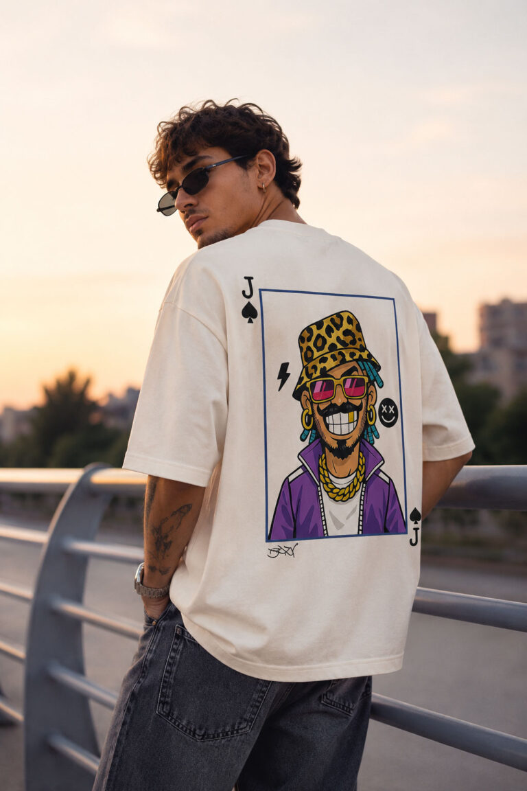 Back view of a man wearing a cream oversized streetwear t-shirt featuring a large cartoon graphic of a character with a gold tooth, leopard bucket hat, and purple jacket inside a playing card frame.