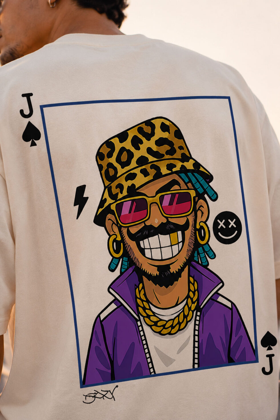 Close-up detail of the vibrant graphic print on the back of a cream heavyweight cotton t-shirt, showing an urban cartoon character with a single gold tooth smile, pink sunglasses, and gold chains.