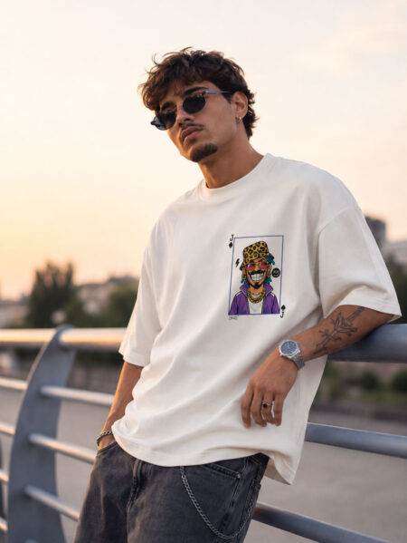 Front view of a man leaning on a bridge railing at sunset, wearing an off-white boxy-fit graphic tee with a small playing card character print on the left chest.