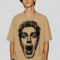 Front view of a male model wearing a beige oversized graphic streetwear t-shirt featuring a large black halftone portrait of a shocked face with an open mouth